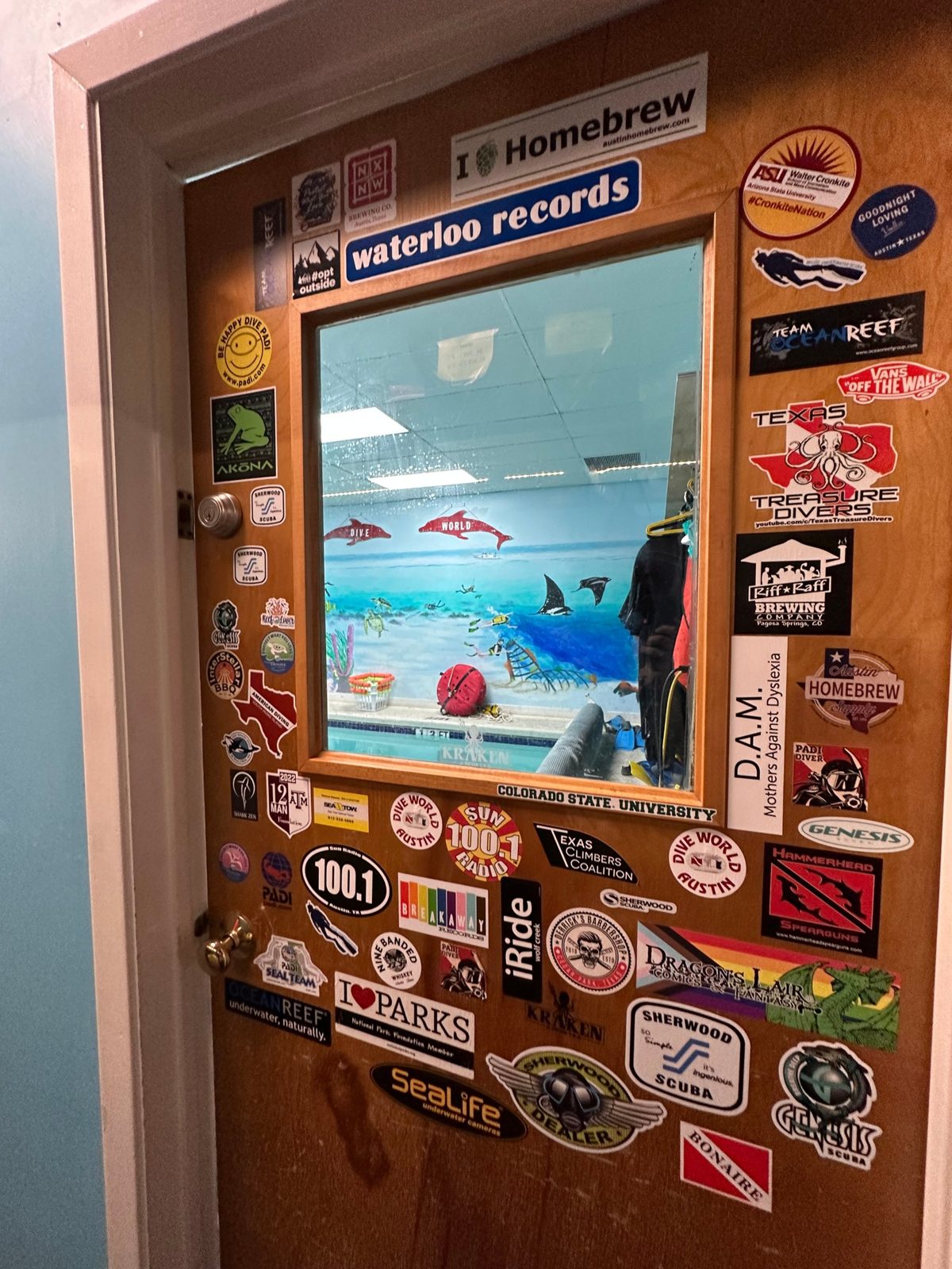 The door to the Dive World Austin pool, covered in stickers, with a window onto the pool and its ocean mural.