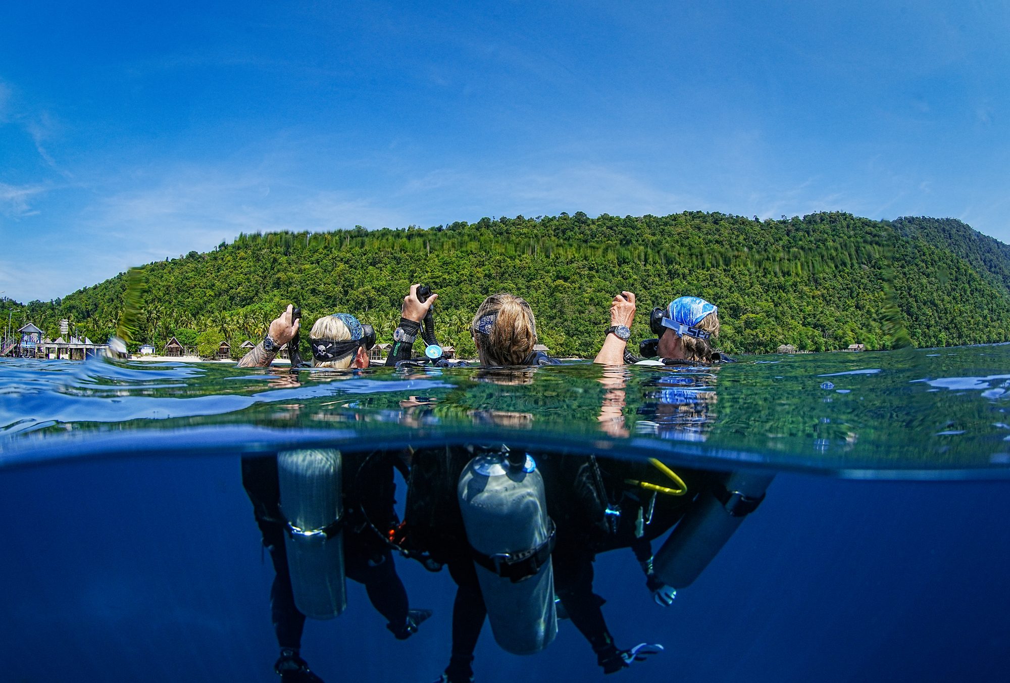 Three divers at the surface, seen from half underwater, about to descend with tropical green hills behind them.