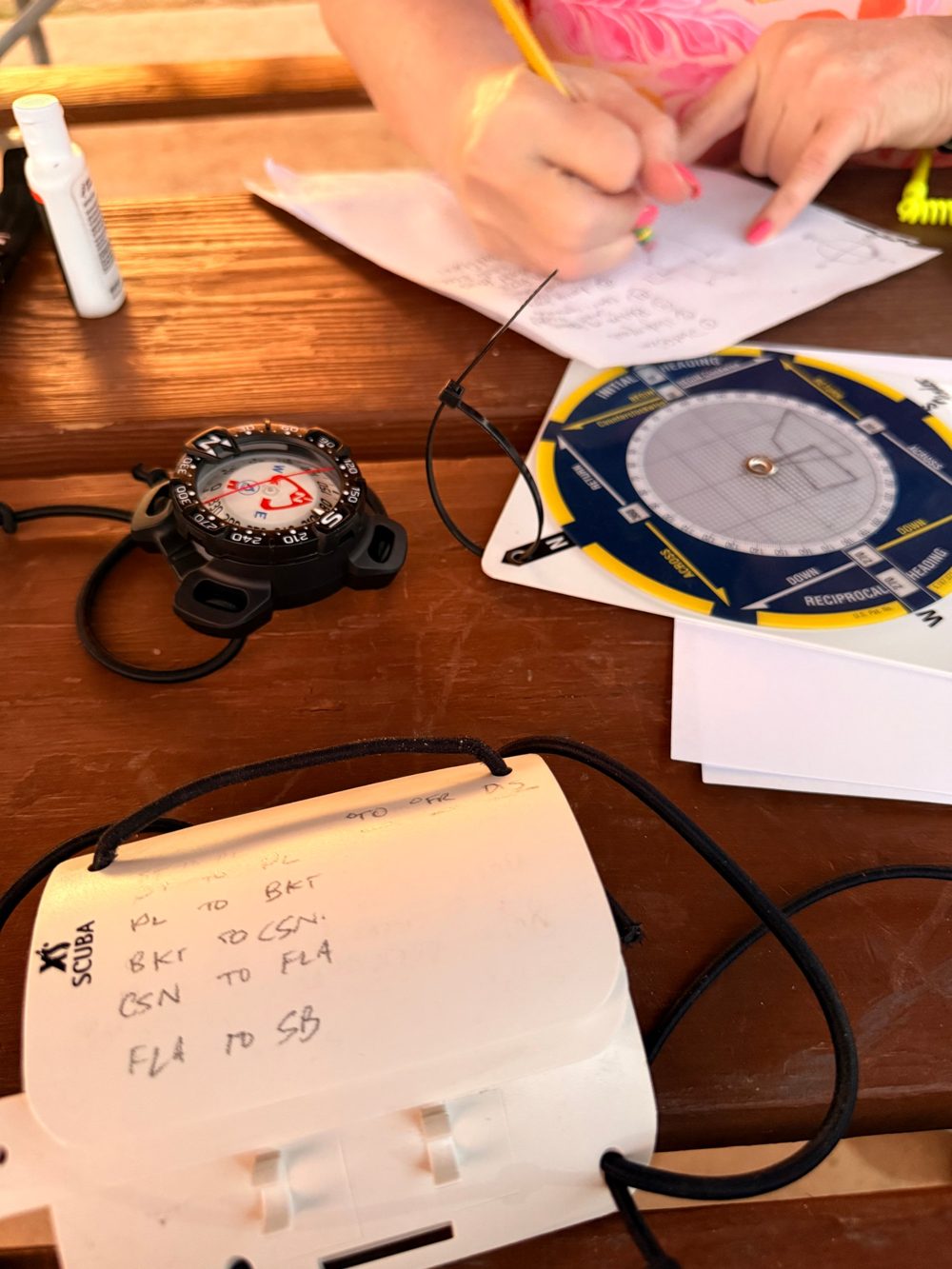 A dive compass and a navigation slate on a picnic table while a student sketches a compass course on paper.