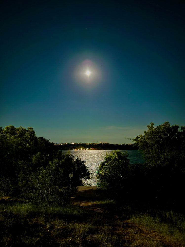 A full moon over a lake at night, its light shining across the water through a gap in the trees.