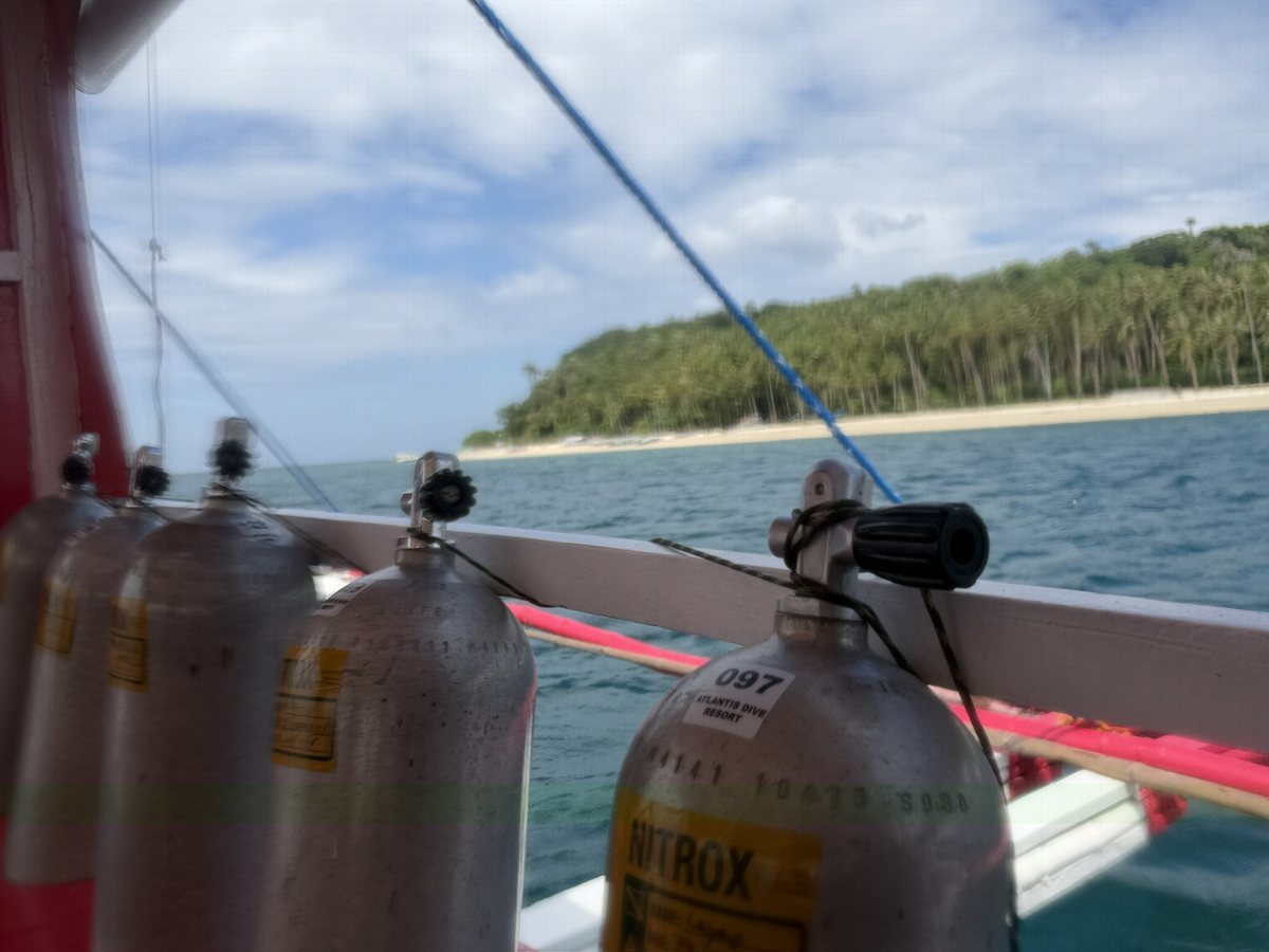A row of scuba tanks on a dive boat rail, the nearest labeled Nitrox, with a tropical island and beach behind.