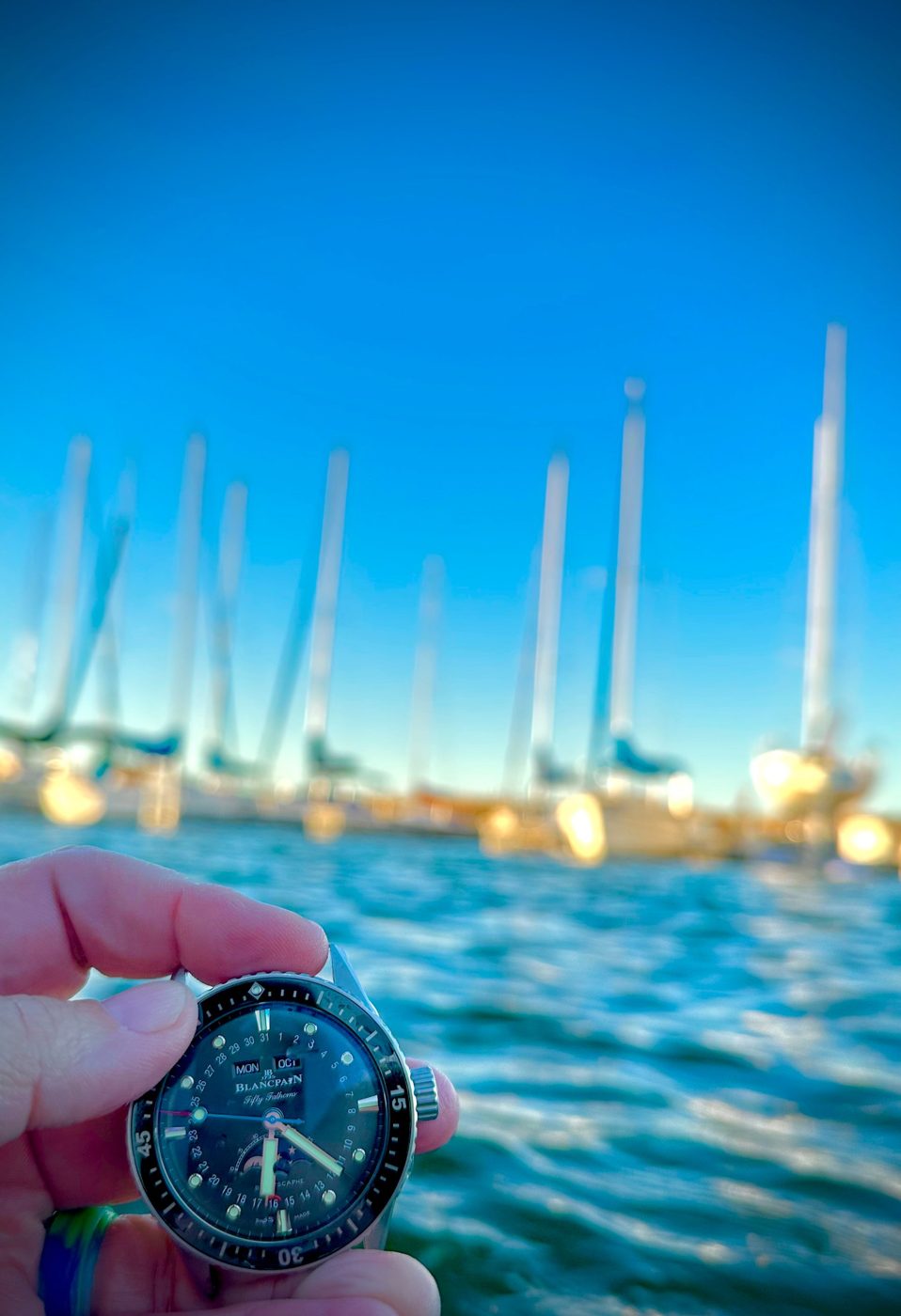 A dive watch held up in front of sailboat masts and choppy blue water.